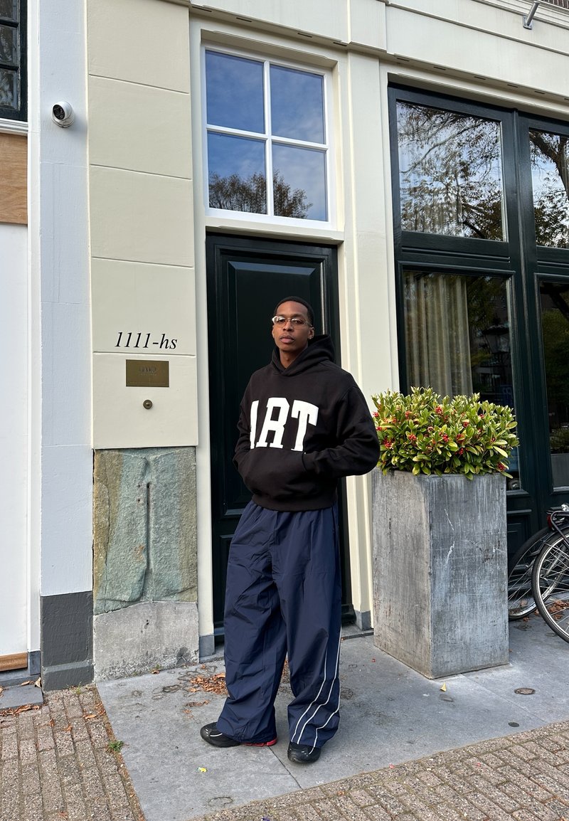 Sweatshirt preto com letras brancas "ART", calças baggy azul-marinho, de pé ao lado de uma porta verde, com um vaso de pedra texturizado e uma bicicleta por perto.