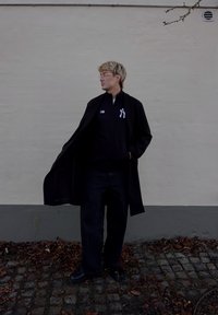 Černý dlouhý kabát přes černou bundu na zip s bílým logem, v kombinaci s tmavými džínami a černými botami, na světlém pozadí stěny.