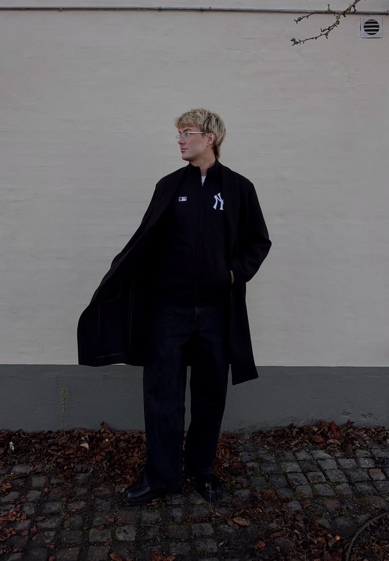 Cappotto lungo nero sopra una giacca nera con zip e logo bianco, abbinati a jeans scuri e scarpe nere, su uno sfondo di muro chiaro.