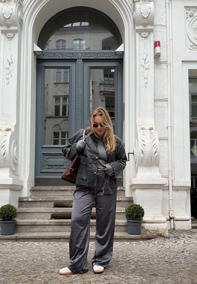 Chaqueta negra sobre suéter gris y pantalones de satén grises, complementados con un bolso marrón y zapatillas blancas, frente a un edificio histórico.