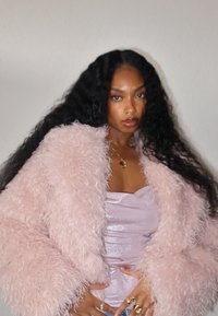 Kobieta z długimi, kręconymi czarnymi włosami nosi jasnoróżową, puszystą kurtkę na błyszczącym lawendowym topie, uzupełnioną złotymi pierścieniami i naszyjnikiem.