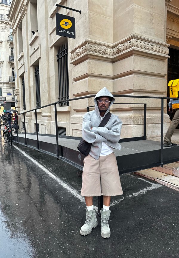 Grijze hoodie, beige korte broek en grijze veterschoenen. Stedelijke omgeving met een helling en een "La Poste"-bord op de achtergrond.