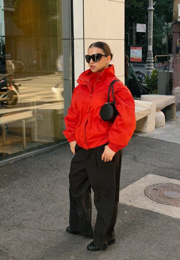 Mujer con gafas de sol oscuras, vistiendo una chaqueta roja brillante y pantalones anchos negros, está de pie en la acera de una ciudad cerca de ventanas de cristal.
