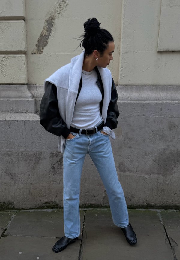 Personne les mains dans les poches porte une veste en cuir noire, un pull clair, un jean bleu clair, des chaussures noires et se tient contre un mur de pierre usé.