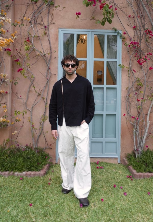 Maglietta nera a maniche lunghe con colletto, pantaloni bianchi a gamba larga e scarpe nere. Sfondo floreale e finestra turchese accentuano la scena.