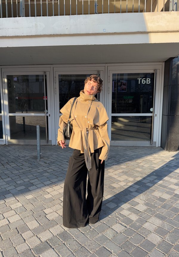 Persoon staand op een verharde ondergrond, draagt een beige jas met riem en wijde zwarte broek, zwarte schoudertas, voor glazen deuren gemarkeerd met T6B.