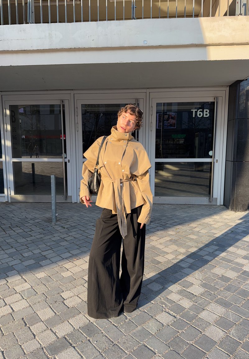 Persona in piedi su un'area lastricata che indossa una giacca beige con cintura e pantaloni neri larghi, borsa nera a tracolla, di fronte a porte di vetro contrassegnate T6B.