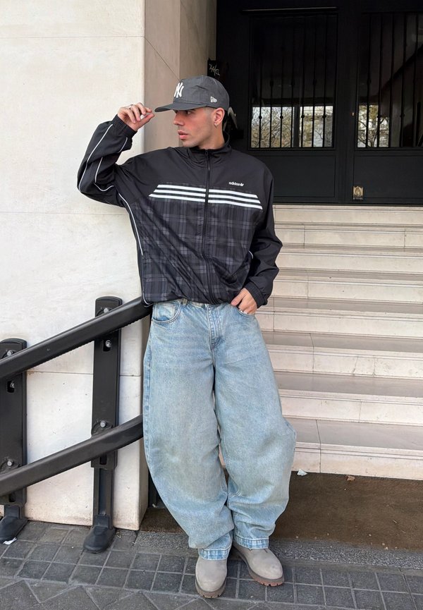 Jonge man in een zwarte Adidas-jas, losse lichtblauwe spijkerbroek, beige laarzen en een grijze New York Yankees-pet poseert bij buitentrappen en leuning.