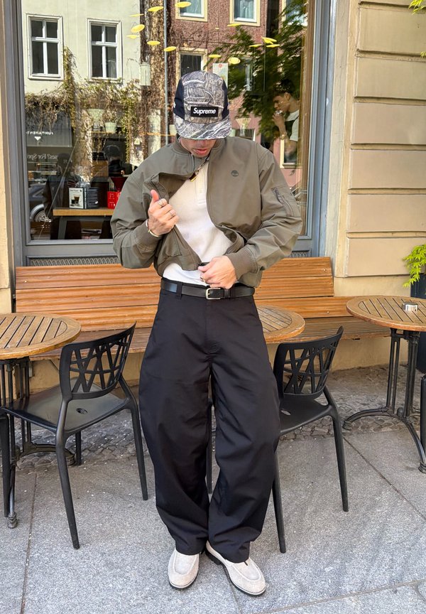 Joven ajustándose una chaqueta marrón, con pantalones anchos negros, zapatos blancos y gorra Supreme gris con camuflaje, de pie fuera de una cafetería con bancos y mesas de madera.