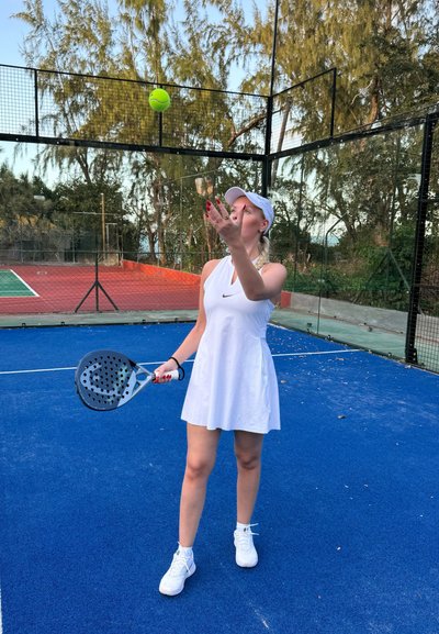 Vestido de tenis blanco, pantalones cortos texturizados y zapatillas blancas. Sosteniendo un remos de pádel. Una pelota de tenis verde está en el aire sobre la mano. Superficie de la cancha azul.