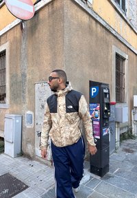 Beige and black The North Face jacket featuring a camouflage pattern, paired with dark blue track pants. An urban environment with a parking meter in close proximity.