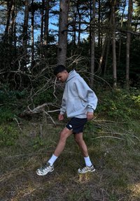 Mikini cinza com um bolso frontal, calções pretos e sapatilhas de corrida com detalhes verdes. Aposentado numa área arborizada com relva seca e ramos.