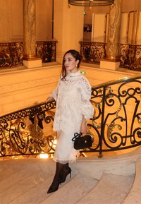 Blondebluse i hvidt blonde med ballonærmer og høj hals, kombineret med en hvid satin nederdel og sorte stiletter. Lille sort håndtaske.