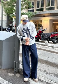 Grijze trui met marineblauwe NY-logo, brede blauwe jeans en beige schoenen. Geaccentueerd met een beige pet en zilveren sieraden. Stedelijke straatomgeving.