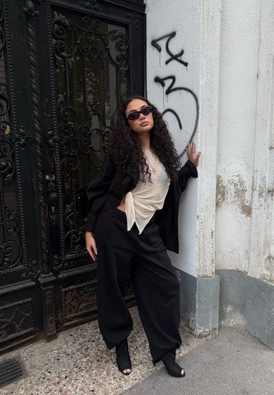 Pantalones amplios negros y una blusa ligera texturizada con un lazo en la parte delantera. Acompañados de una chaqueta corta negra y unas pequeñas gafas de sol negras.