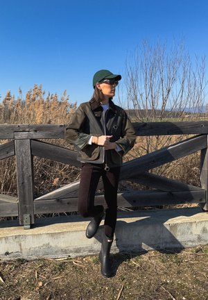 Persona con chaqueta oscura, pantalones negros, botas negras, gorra verde y gafas de sol, de pie junto a una cerca de madera con plantas secas y cielo azul claro de fondo.