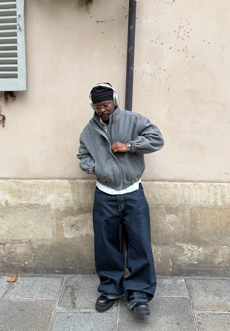 Veste bomber grise zippée, t-shirt blanc en dessous, jean décontracté en denim foncé, et bottes noires épaisses. Personne portant un casque audio et des lunettes.