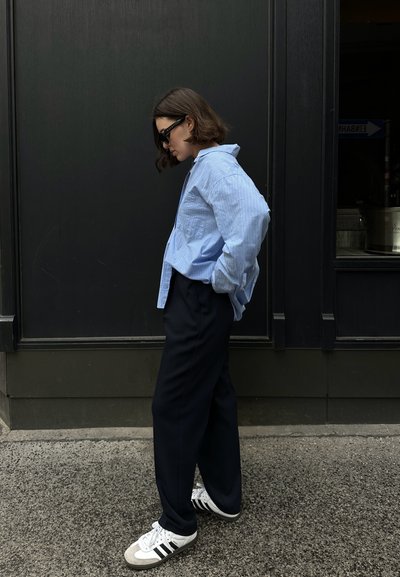 Camisa de botones a rayas azul claro, pantalones negros holgados y zapatillas blancas con rayas negras. De pie contra una pared oscura.