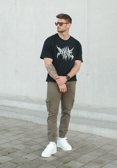 Hombre con gafas de sol, camiseta negra con estampado gráfico, pantalones cargo oliva, zapatillas blancas y reloj, de pie con las manos entrelazadas delante sobre el pavimento.