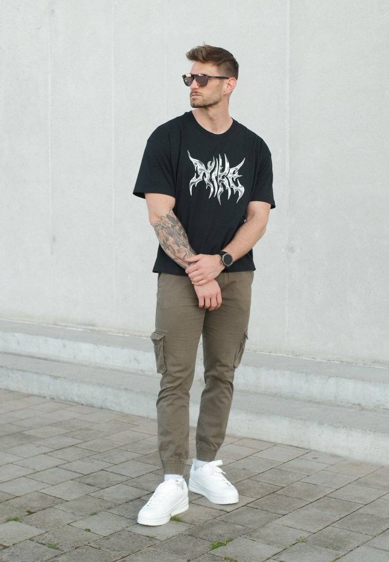 Homme portant des lunettes de soleil, un t-shirt noir avec un motif graphique, un pantalon cargo olive, des baskets blanches et une montre, debout les mains jointes devant sur le trottoir.
