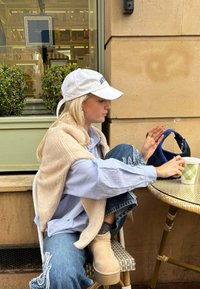 Μπεζ oversized γιλέκο από μάλλινο ύφασμα, τοποθετημένο πάνω από πουκάμισο σε μπλέ καρό σχέδιο, σκισμένα τζιν, μπεζ πλατφόρμες, καθισμένος σε ένα τραπέζι καφετέριας.