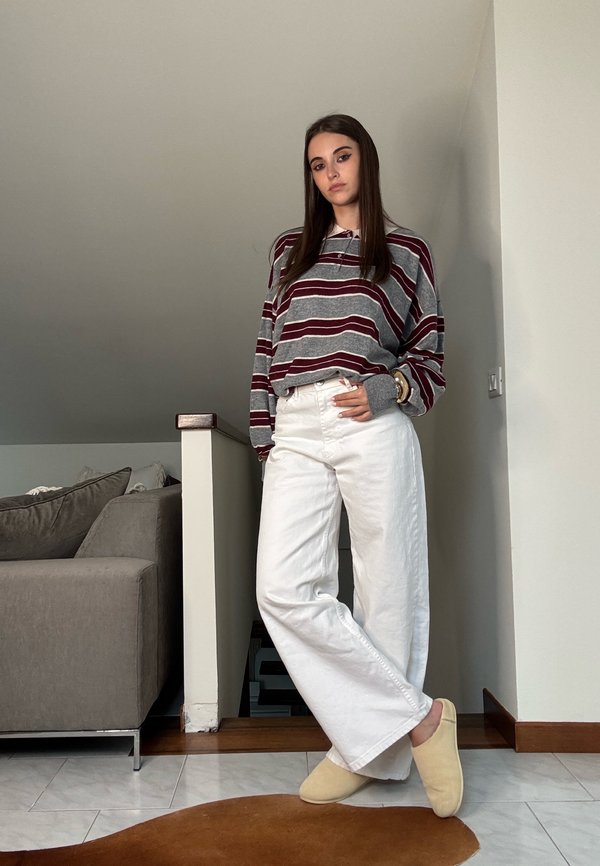 Striped grey and burgundy long-sleeve shirt, white wide-leg trousers, beige slippers, standing indoors with a neutral backdrop and soft furnishings.