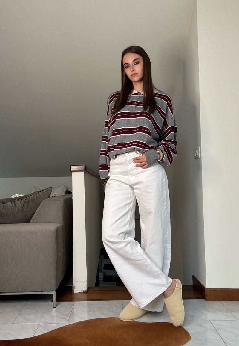 Gestreepte grijs en bordeauxrode blouse met lange mouwen, witte wijde broek, beige slippers, staand binnen met een neutrale achtergrond en zachte meubels.