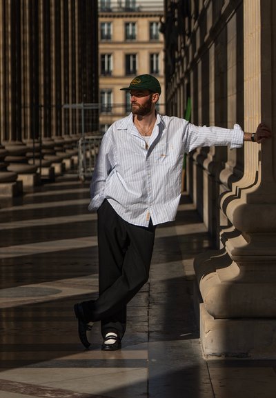 Camisa de botones blanca y azul a rayas, pantalones negros, zapatos negros con acentos blancos, gorra verde, de pie contra una columna de piedra.