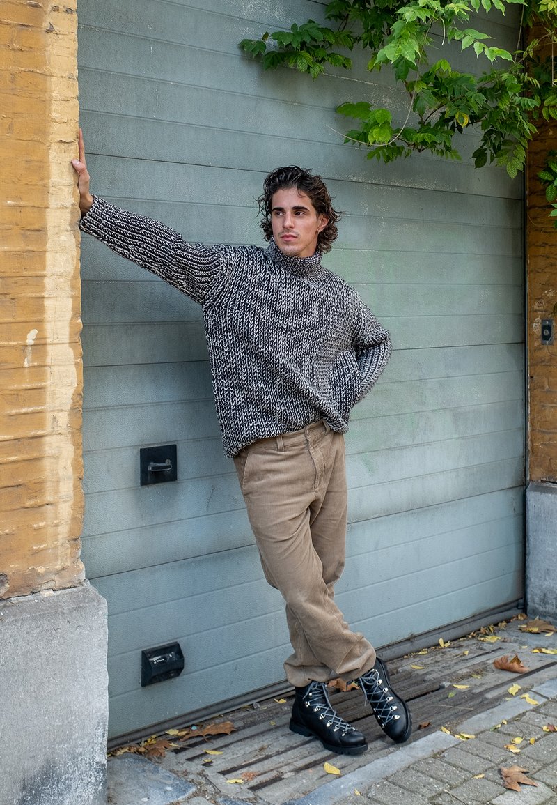 Melnai ir pilkų atspalvių megztinis, aukštu kaklu, derinamas su smėlio spalvos kordūro kelnėmis ir juodais raišteliais batais, prieš tekstūruotą sieną.