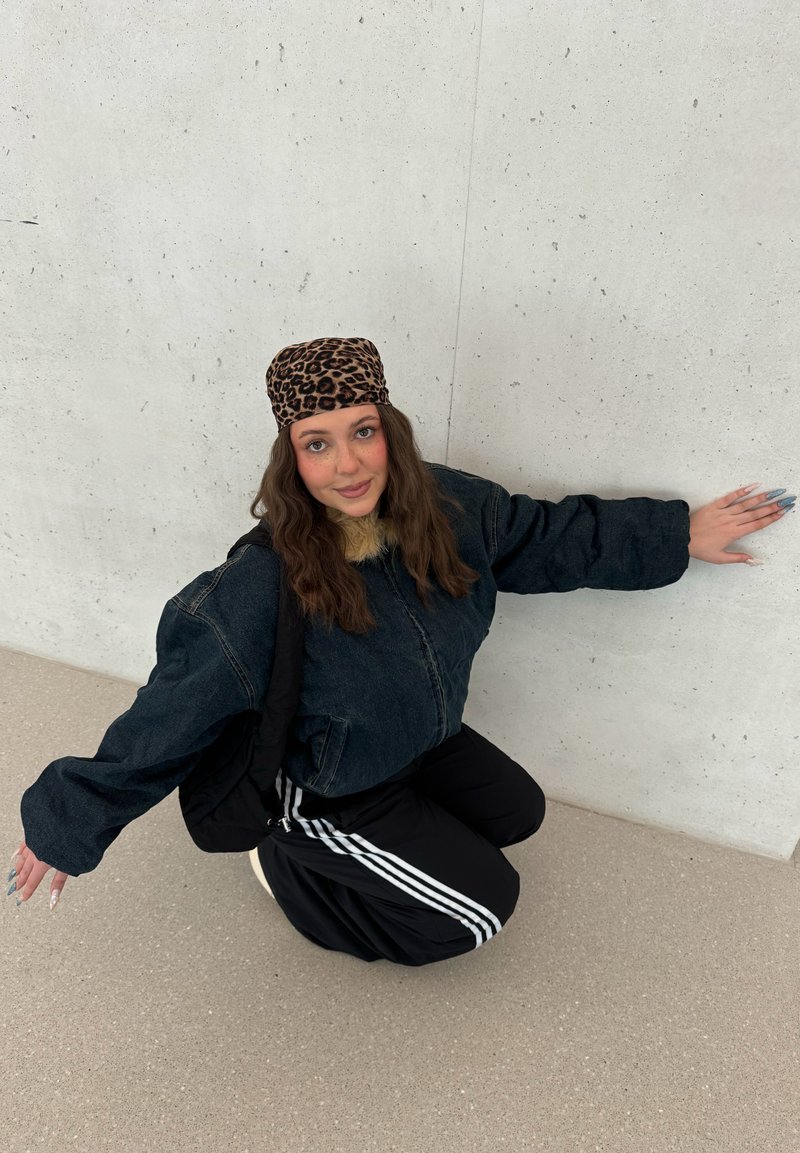 Jeune femme en veste sombre, foulard à imprimé léopard et pantalon noir avec des bandes blanches, posant à genoux contre un mur de béton clair.