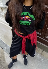 Svart sweatshirt med tecknad grafik och grön text, kombinerad med svarta shorts. Röd- och svartrandig flanell skjorta knuten runt midjan. Svarta sneakers.