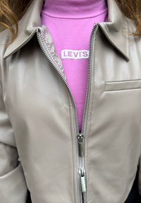 Beige läderjacka med silverfärgade dragkedjedetaljer, med en liten bröstficka, bärs över en rosa polotröja med en vit Levi's-logga.