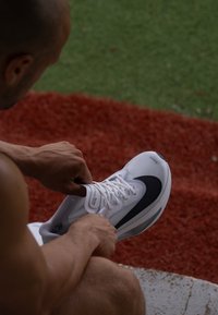 Hvit joggesko med mesh-overdel, sorte detaljer og grå såle; bli snøret av en person som sitter på en grusoverflate.