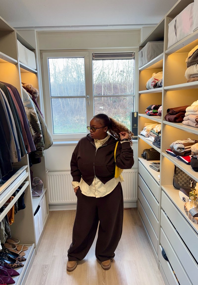 Personne portant une tenue marron foncé et des lunettes se tient dans un dressing lumineux avec des étagères de vêtements pliés et des vestes suspendues.