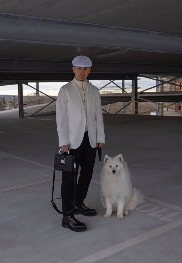 Vit blazer över en ribbad tröja, svarta byxor och kraftiga svarta stövlar. Håller en svart väska; fluffig vit hund sitter bredvid honom i ett parkeringsgarage.