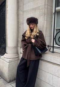 Manteau en laine marron à manches larges et ceinture, associé à un pantalon noir à jambes larges et un sac à main en cuir noir. Un chapeau en fourrure complète le look.