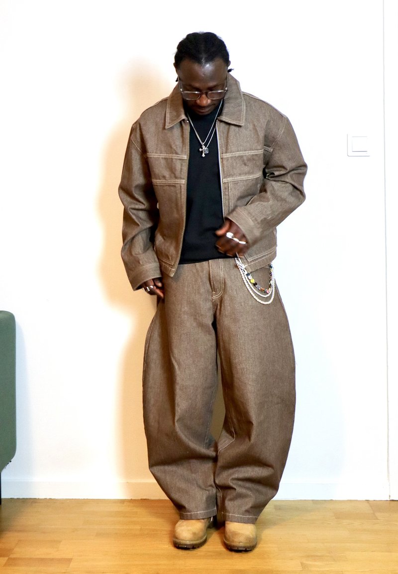 Man wearing a brown oversized denim jacket and trousers, black shirt, tan boots, glasses, and layered necklaces, standing indoors on a wooden floor.