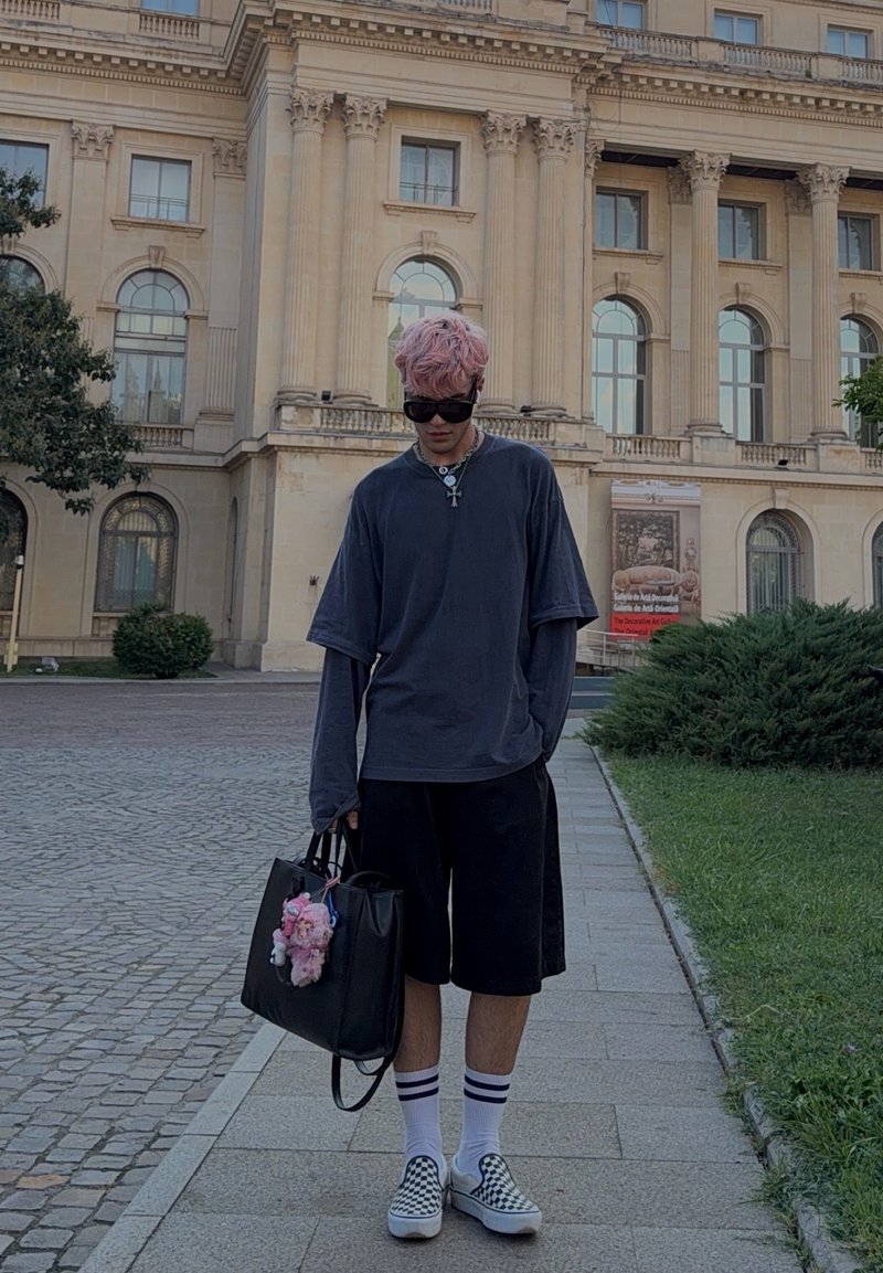 Jeune personne aux cheveux roses et lunettes de soleil, portant des vêtements sombres et amples, tenant un sac noir avec des peluches, debout sur un trottoir en ville.