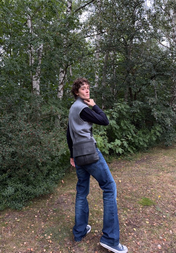 Person med kortt krøllet hår iført grå vest og jeans står i en lysning i skoven og kigger over skulderen.