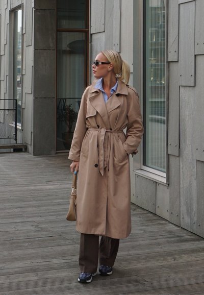 Abrigo beige de trench con cintura ceñida, combinado con pantalones marrones y zapatillas deportivas grises. Se ven gafas de sol y una camisa azul clara.