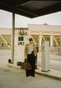 En man i en beige trenchcoat och svart hatt står vid vintage bensinpumpar. Han håller en brun rutig duffelväska och har på sig svarta byxor.