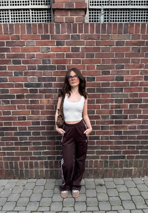 Pantalones deportivos burdeos con rayas blancas, top corto ajustado blanco y bolso marrón. El fondo presenta una pared de ladrillos y pavimento gris.