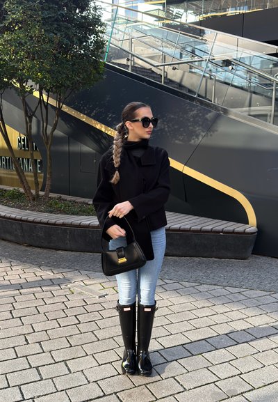 Abrigo negro recortado sobre un suéter de cuello alto negro, jeans azules y botas de lluvia negras brillantes hasta la rodilla. Sostiene un bolso negro con detalles dorados.