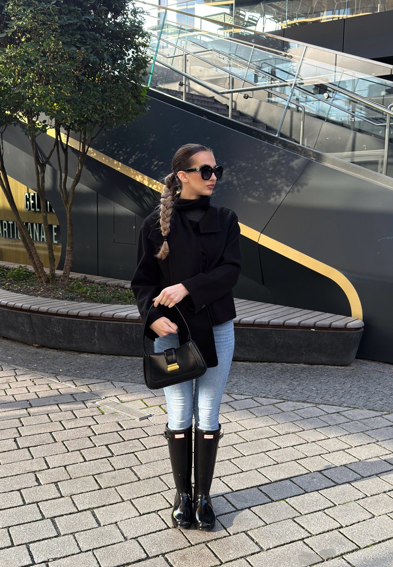 Casaco preto curto sobre uma camisola de gola alta preta, jeans azuis e botas de chuva pretas brilhantes até ao joelho. Segura uma mala preta com ferragens douradas.