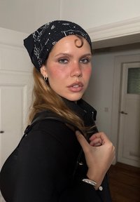 Czarna bandana w wzory z białymi motywami niebieskimi, kontrastująca z jasnobrązowymi włosami i czarną kurtką. Akcenty w postaci srebrnych kolczyków i bransoletki.