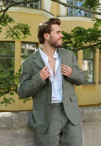 Traje de lino verde claro con chaqueta estructurada y pantalones a juego, combinado con una camisa blanca y un collar de cadena de plata.