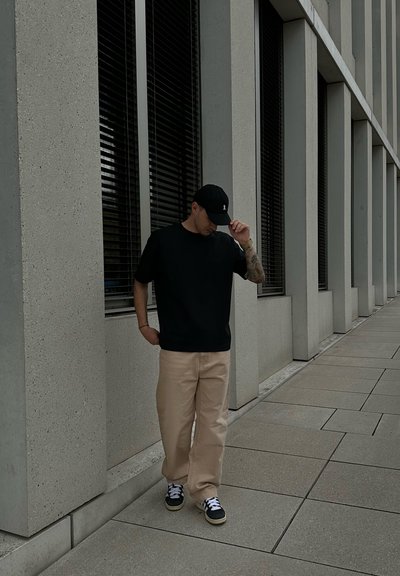 Camiseta negra, pantalones baggy beige, gorra negra con logo, zapatillas negras y blancas con patrón; de pie frente a un fondo de edificio de concreto.