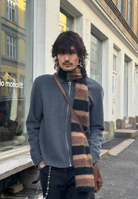 Veste grise zippée en tissu texturé, écharpe multicolore à rayures et pantalon noir. Les détails incluent un sac à sangle marron et des bijoux.