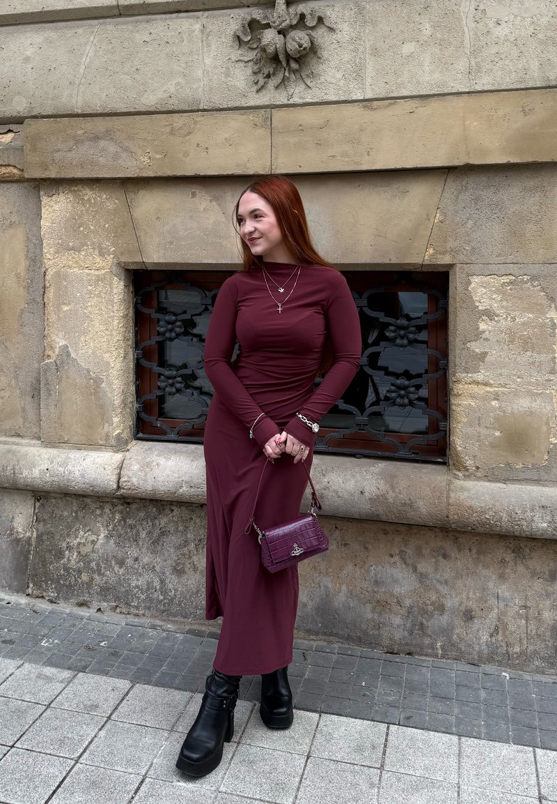 Longue robe bordeaux à manches longues, assortie d'un petit sac à main et de bottines à plateforme noires, devant un mur en pierre texturé.