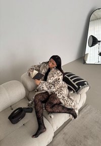 Leopardmönstrad faux fur-kappa, svarta spetsstrumpbyxor, sittande på en krämfärgad soffa med mönstrad kudde, läser en bok. Svart handväska i närheten.